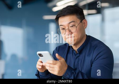 Primo piano. Un giovane asiatico arrabbiato in occhiali sta tenendo un telefono nelle sue mani. Nervosamente le labbra pursed, legge notizie, messaggi, perde nei giochi online. Foto Stock