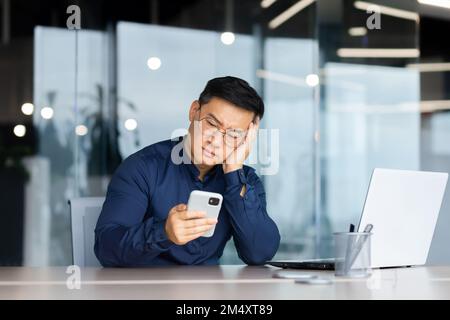 Ritratto di un giovane asiatico preoccupato seduto in ufficio al tavolo, tenendo la testa con la mano, guardando tristemente il telefono. Annoiato, ricevuto cattive notizie, messaggi. Foto Stock