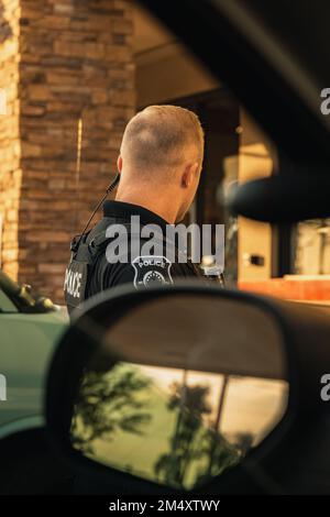 Immagine verticale di un poliziotto caucasico maschio bianco in piedi fuori dal veicolo poliziotto visto dall'interno della macchina da trouper. Ufficiale che guarda lontano da venuto Foto Stock