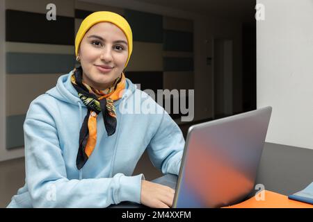 Sorridente ragazza in una felpa blu e cappello giallo funziona su un computer nel corridoio Foto Stock