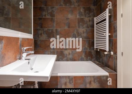 Bagno con lavabo a parete in porcellana bianca senza armadio, specchio con cornice in legno bianco, piastrelle a chiazze e portasciugamani bianco Foto Stock