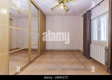 Camera vuota con armadio da muro a muro con porte scorrevoli in rovere e ventilatore a soffitto a specchio, pareti dipinte in crema e pavimento piastrellato marrone chiaro e ba Foto Stock