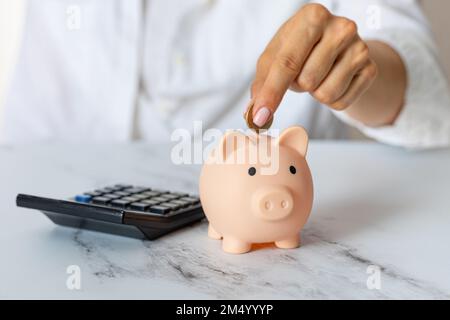 Primo piano delle mani di una donna mettere una moneta in euro in una banca piggy, accanto a una calcolatrice sul tavolo.investimento corretto. Risparmio di denaro in tempi di crisi e. Foto Stock