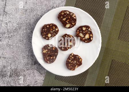 Salsiccia di cioccolato fatta in casa. Il dessert a base di biscotti, cioccolato e noci viene affettato su un piatto rotondo bianco su sfondo scuro. Vista dall'alto, disposizione piatta Foto Stock