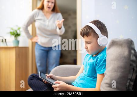 Madre scolda suo figlio. Il ragazzo usa il tablet con le cuffie e ignora la mamma Foto Stock