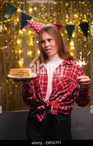 Foto di giovane infelice triste sconvolto dispiacere, mostra lingua, con torta di compleanno in festa nel salotto decorato a casa. Spazio di copia Foto Stock