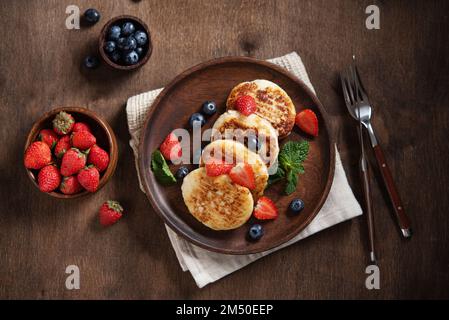 Frittelle al formaggio o sciroppi con mirtilli, fragole e menta fresca su uno sfondo di legno scuro. Il concetto di una pausa sana fatta in casa Foto Stock