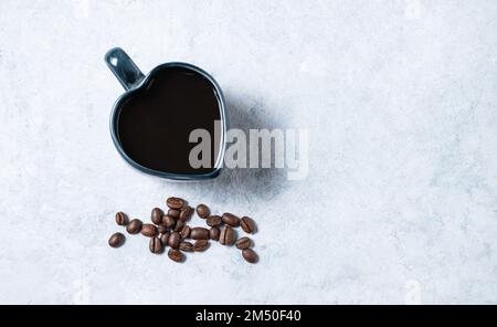 Una tazza di caffè espresso nero a forma di cuore con grani sparsi su sfondo blu. Vista dall'alto e spazio di copia. Foto Stock