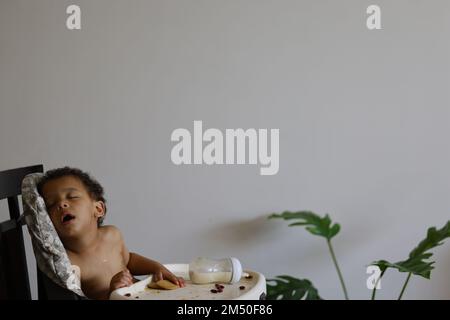 Un ragazzino carino che si è addormentato nel seggiolone dopo aver mangiato il pasto Foto Stock