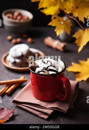 Tazza rossa di cioccolato caldo aromatico con marshmallows su uno sfondo marrone con foglie di acero autunnale, cannella e biscotti. Concetto accogliente drink. Vista frontale Foto Stock