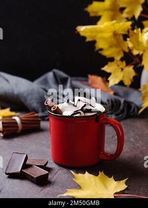 Tazza rossa di aroma cioccolata calda con marshmallows su fondo marrone con foglie di acero autunnale e sciarpa grigia. Concetto accogliente drink. Vista frontale e cl Foto Stock