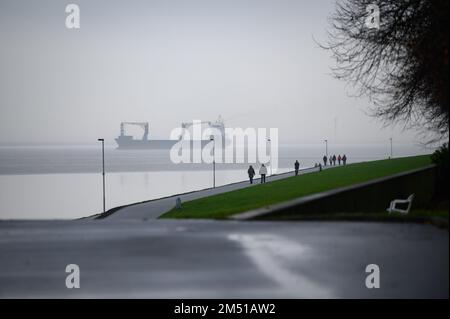 24 dicembre 2022, Schleswig-Holstein, Brunsbüttel: Gli escursionisti camminano lungo una diga mentre una nave container naviga nella nebbia sull'Elba. Foto: Jonas Walzberg/dpa Credit: dpa Picture Alliance/Alamy Live News Foto Stock