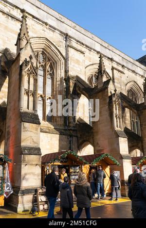 Gli amanti dello shopping natalizio al mercatino di Natale di Winchester con bancarelle tra i contrafforti volanti in pietra della cattedrale gotica di Winchester, Inghilterra, Regno Unito Foto Stock