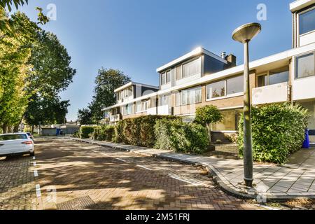 una strada con auto parcheggiate di lato e alberi che costeggiano la strada di fronte all'edificio, che è circondato da cespugli Foto Stock