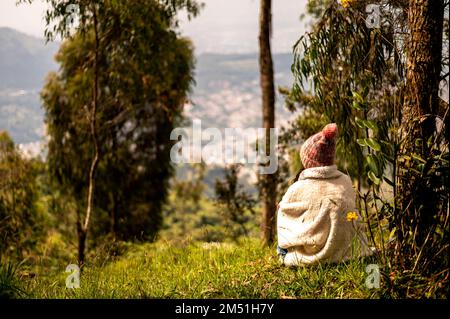 Donna fredda vestita di poncho e cappello seduto in collina erbosa, prendendo il sole e ammirando la vista Foto Stock