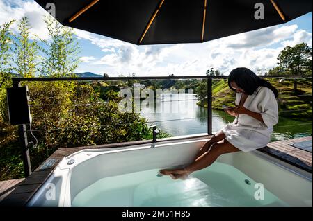 donna vestita di ruba si siede sul bordo di una jacuzzi all'aperto con le sue feste nella jacuzzi guardando il suo telefono Foto Stock