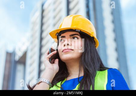 giovane ingegnere caucasico venezuelano donna, indossando casco giallo di sicurezza e giubbotto riflettente, in piedi in strada per una telefonata, ascoltare e thi Foto Stock