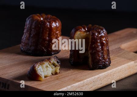 Un primo piano di Caneles e una fetta perfettamente tagliata su un tagliere di legno e sfondo scuro Foto Stock