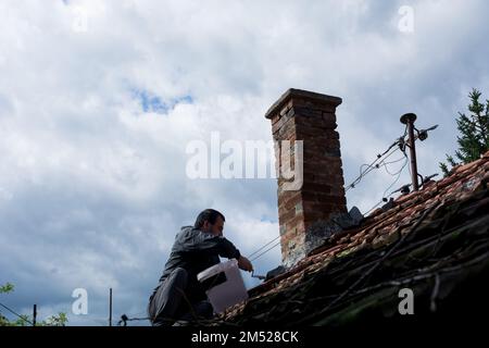 Un artigiano sul tetto di una vecchia casa che fissa il camino con una cazzuola. Conservando la struttura, si siede in una tranquilla scena di duro lavoro Foto Stock