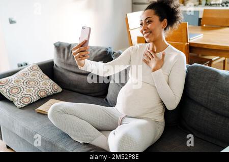 Giovane bella sorridente felice donna afro incinta seduta sul divano toccando il petto e prendendo selfie al telefono in una stanza accogliente a casa Foto Stock