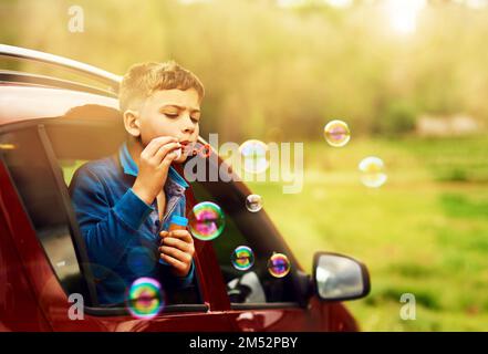 Le bolle perfette richiedono messa a fuoco e tecnica. un ragazzino giocoso che soffia delle bolle mentre si inclina fuori dal finestrino di un'auto. Foto Stock