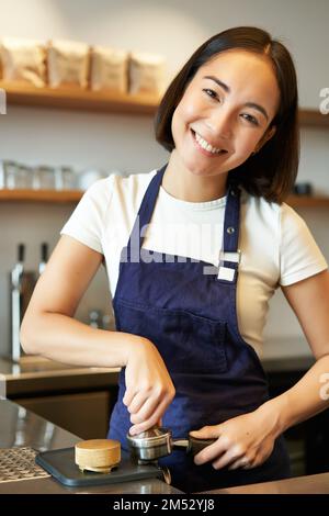 Scatto verticale di carino barista asiatico, ragazza sorridente mette il caffè in manomissione, preparare il cappuccino, in piedi al banco del caffè Foto Stock