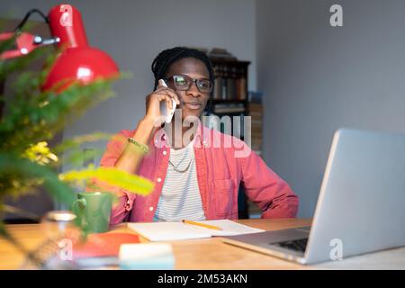 Un ragazzo africano sorridente che parla al telefono mentre si siede alla scrivania con un computer portatile, distratto dallo studio online Foto Stock