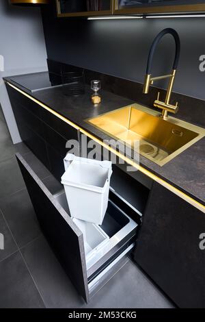 Armadio del cestino della cucina. Estrarre il cassetto della cucina per il cestino raccolta differenziata sotto il lavabo dorato e il rubinetto piano cottura elettrico in vetro nero Foto Stock