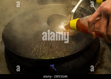 Uno chef asiatico prepara una salsa dolce e acida in un wok per friggere alcune spaghetti con le verdure Foto Stock