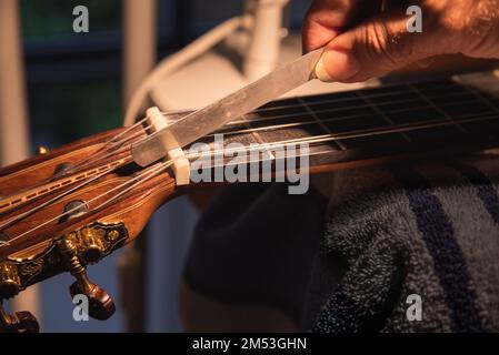 Riparazione e servizio di chitarra per strumenti musicali - la mano operatrice usa il file per riparare il dado osseo della chitarra posto sul collo della chitarra. Foto Stock
