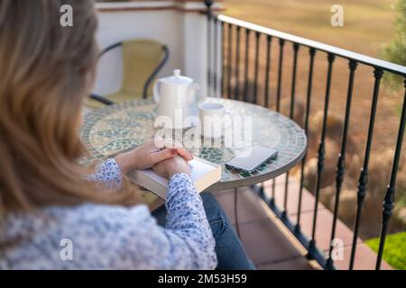 Donna seduta sulla terrazza del suo appartamento con un libro e bere una tazza di caffè in modo rilassato e tranquillo. Foto Stock
