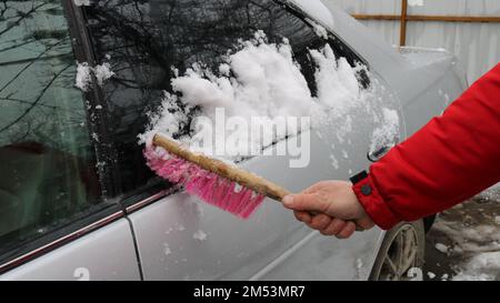 una persona pulisce i depositi di neve sulla superficie di un'auto grigia con una lunga spazzola, pulisce un'auto coperta di neve nel cortile, spazzolando la neve da un veicolo Foto Stock