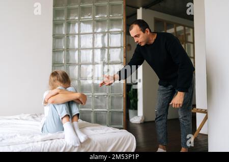 Furioso giovane padre che scola, alzando la voce, urlando e puntando con il dito indice a bambina difficile testardo seduto sul letto Foto Stock