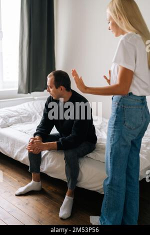 Colpo verticale di giovane marito disperato seduto sul letto guardando via ignorando la moglie bionda arrabbiata litigando l'uomo turbato colpetto di problemi a casa. Foto Stock