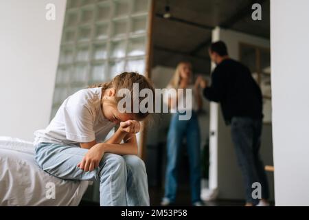 Vista dal basso angolo della disperazione bambina che piange solitaria soffrendo seduto sul divano durante la disputa dei genitori e la lotta in soggiorno sullo sfondo. Foto Stock