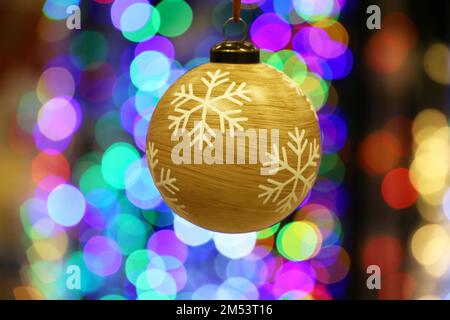 Giocattolo di Capodanno, palla di legno con motivo a fiocchi di neve. Decorazioni natalizie appese su sfondo sfocato luci festive Foto Stock