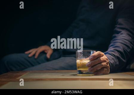 Uomo che tiene un bicchiere di whiskey su un tavolo in condizioni di scarsa illuminazione. Foto Stock