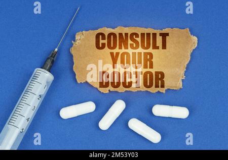 Concetto medico. Su una superficie blu si trovano una siringa, pillole e un cartoncino con l'iscrizione - consultare il medico Foto Stock