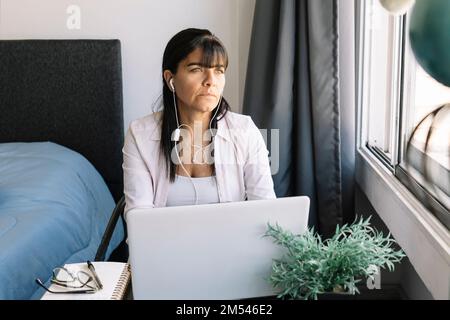 Latina donna che lavora nel suo appartamento guarda verso la finestra. Concetto di home Office, lavoro eccessivo, scadenze e stile di vita. Foto Stock