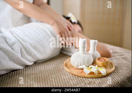 Concetto di spa tailandese, palline di erbe o francobolli da massaggio e sale spa sul tavolo da massaggio con una giovane donna che riceve un massaggio sul tavolo da massaggio. Foto Stock