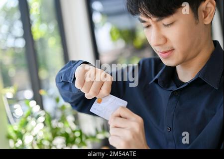 Stressato e arrabbiato giovane uomo asiatico rompere la sua carta di credito, distruggendo la sua carta di credito, avendo problemi finanziari. Foto Stock