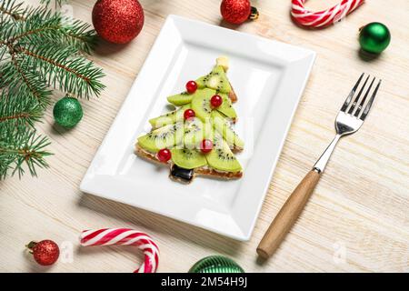 Piatto di gustosa frittella a forma di albero di Natale con frutta, rami di abete e decorazioni su sfondo ligneo Foto Stock