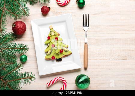 Piatto di gustosa frittella a forma di albero di Natale con frutta, rami di abete e decorazioni su sfondo ligneo Foto Stock