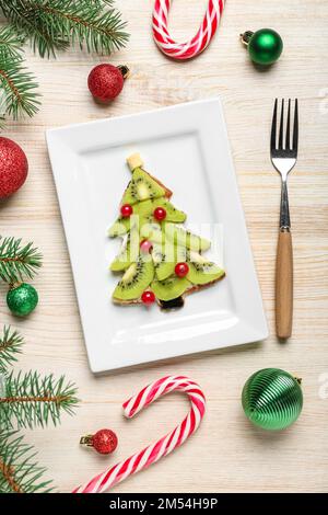 Piatto di gustosa frittella a forma di albero di Natale con frutta, rami di abete e decorazioni su sfondo ligneo Foto Stock