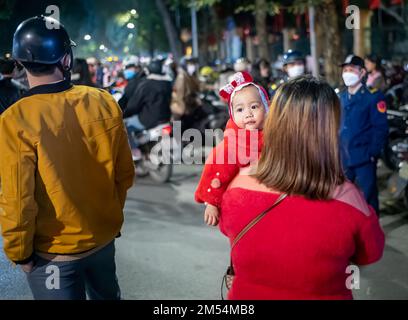 Un bambino vestito di rosso festivo guarda sopra la spalla della madre mentre lo trasporta attraverso la folla nel centro di Hanoi in Vietnam la vigilia di Natale. Foto Stock