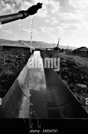 Una verticale di acqua che si versa da un tubo in un tubo metallico in un villaggio sparato in scala di grigi Foto Stock