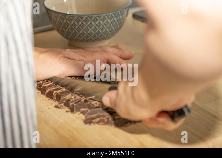 Un primo piano di uno chef che trita la barra di cioccolato in pezzi con un coltello su un tagliere Foto Stock