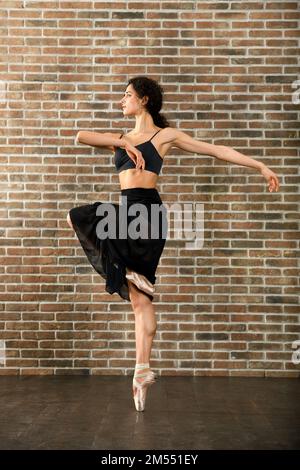 Graziosa ballerina giovane che prova in uno studio posando en pointe di fronte ad un muro di mattoni Foto Stock