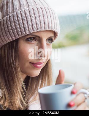 una donna in una cuffia di lana, penosa, che tiene una tazza di caffè. concetto di relax e benessere. Foto Stock