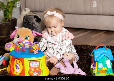 Una ragazza felice di un anno e mezzo gioca con i giocattoli seduti sul pavimento. Foto Stock